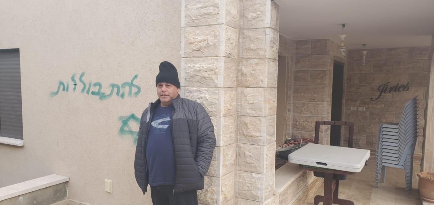 A resident of Jish village stands next to a slogan sprayed by Israeli settlers saying 'Stop assimilating' (Arab48 news)