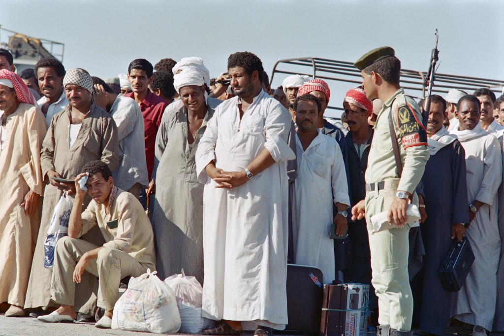 A group of Egyptian men stand together with essentials, including a boxed cassette player, at their feet. 