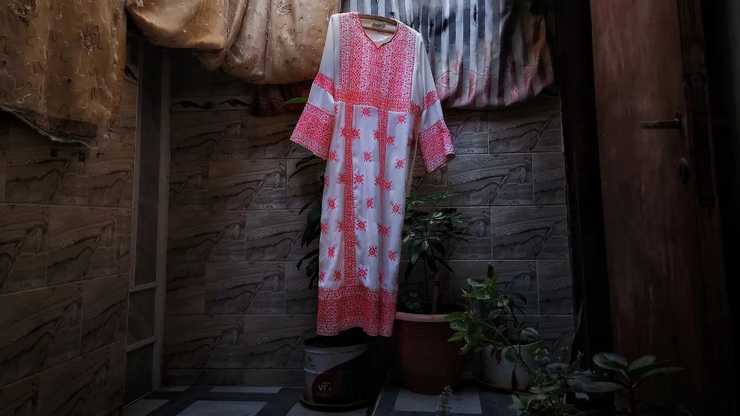 A white dress with red tatreez Palestinian embroidery is hanging on a washing line in a garden.