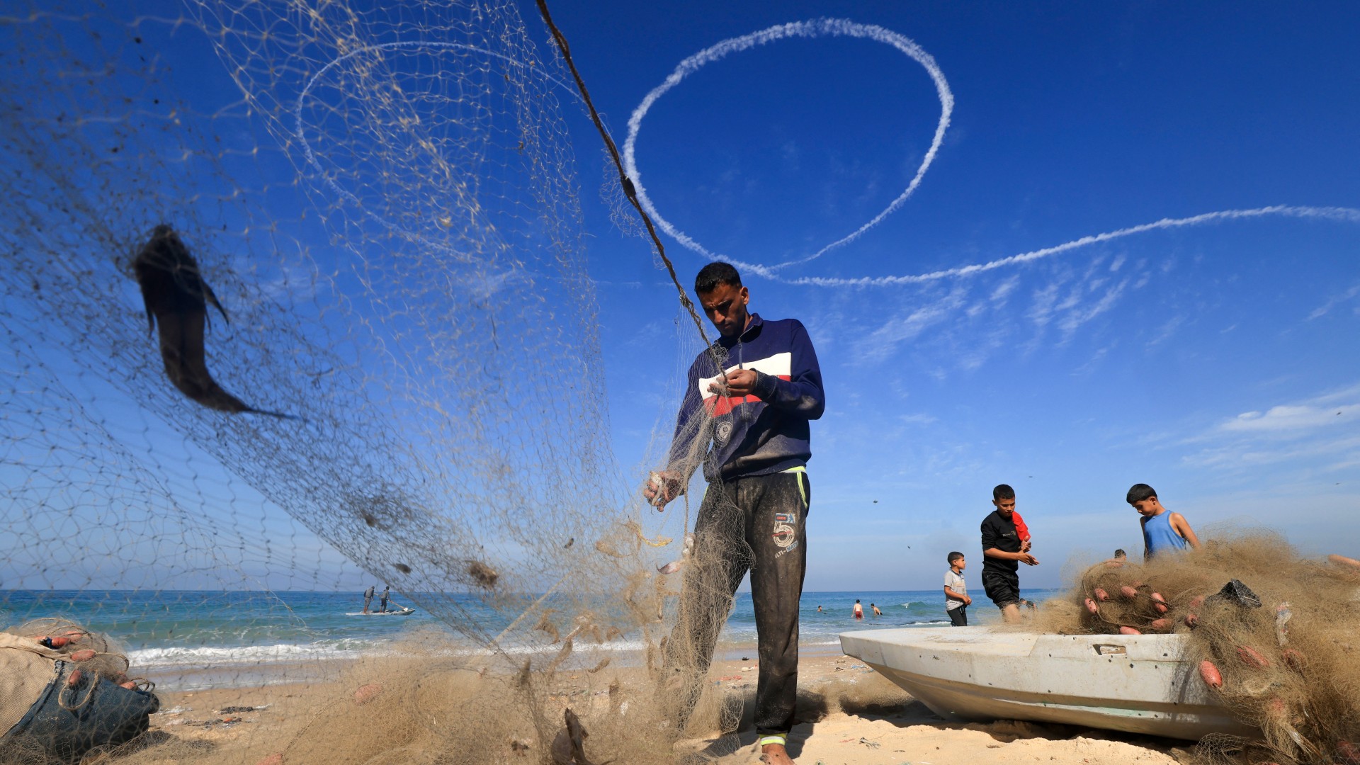 Des pêcheurs palestiniens trient leurs prises à Deir al-Balah alors que les traînées de vapeur d’un avion israélien tapissent le ciel, le 30 novembre (AFP/Mahmud Hams)