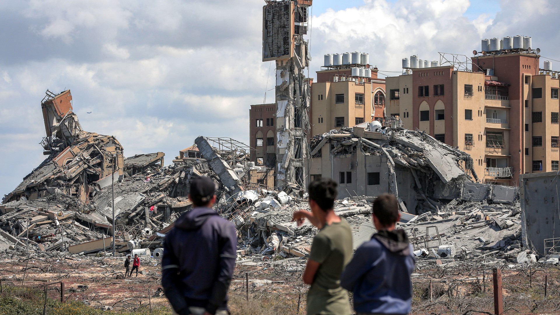 Youths watch from afar as people search the rubble of destroyed buildings in the Asra residential compound, northwest of Nuseirat in the Gaza Strip, on March 25, 2024