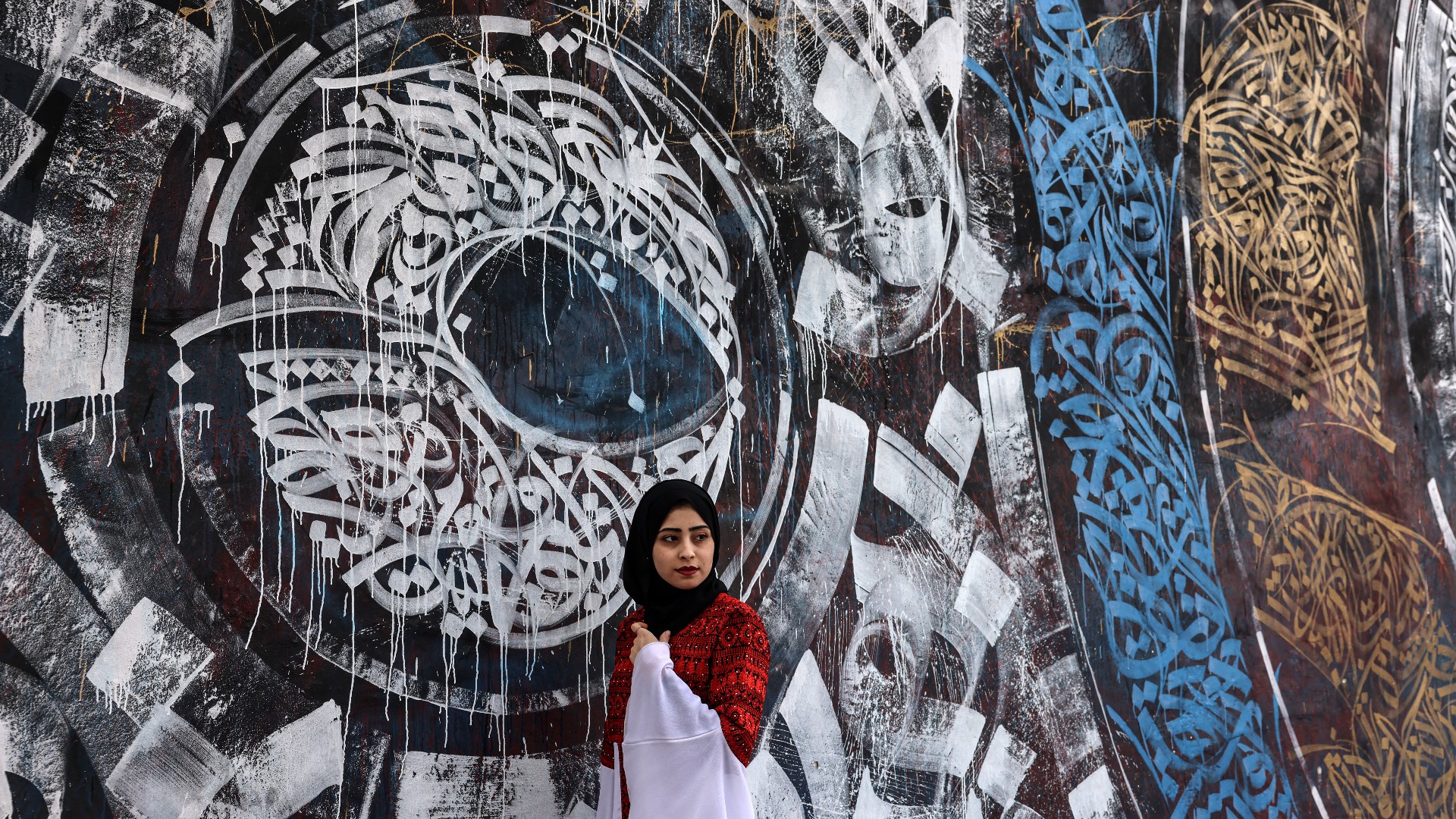 A Palestinian university student wearing a traditional embroidered dress, stands in front of a mural painting representing Arab caligraphy, during an event entitled 'Don't Steal Our Heritage' in Gaza City, on 16 December, 2021 (AFP)