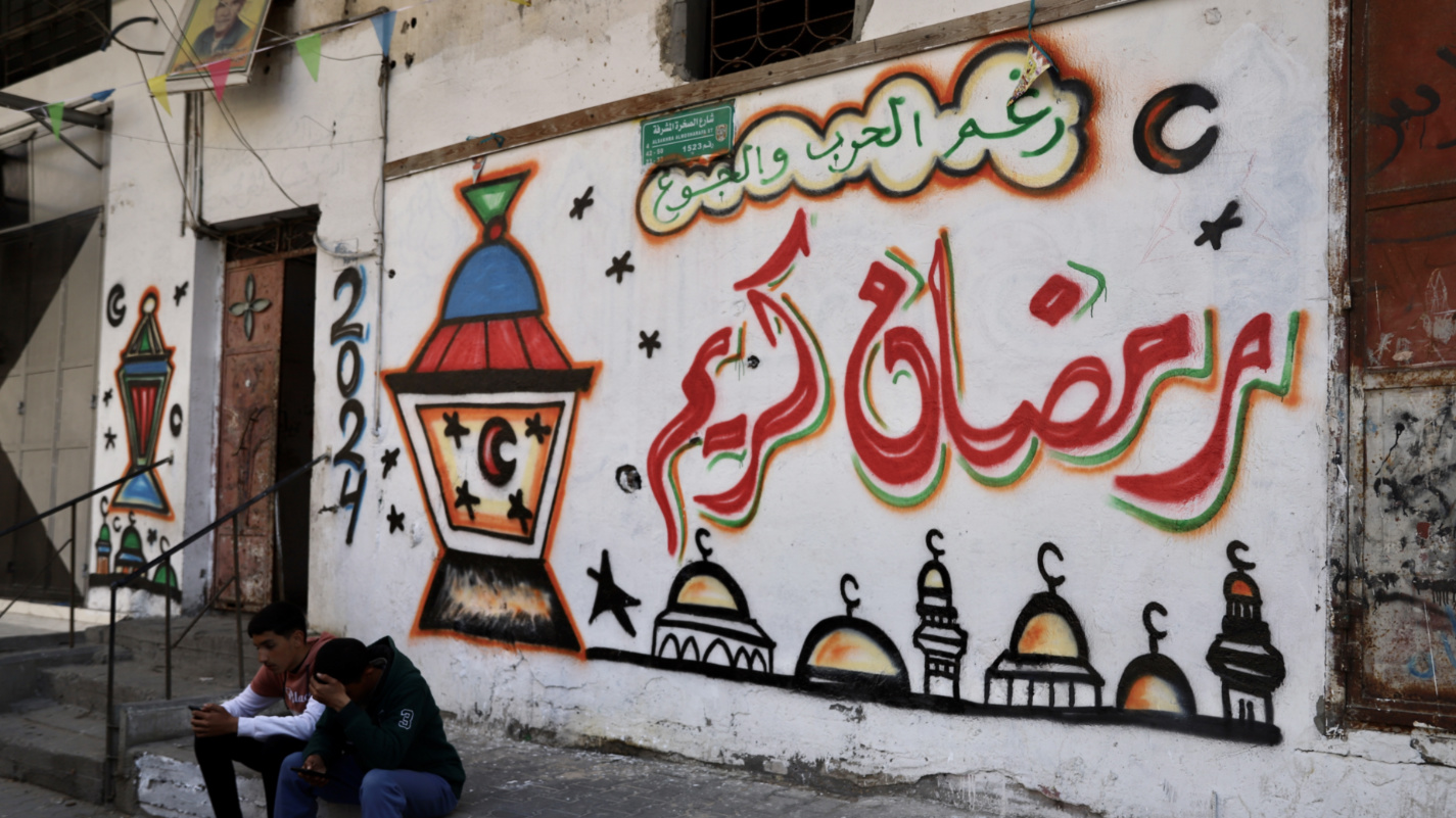 men sit in front of a wall painting in northern Gaza that reads "in spite of war and hunger, have a blessed Ramadan" (Mohammed al-Hajjar/MEE).