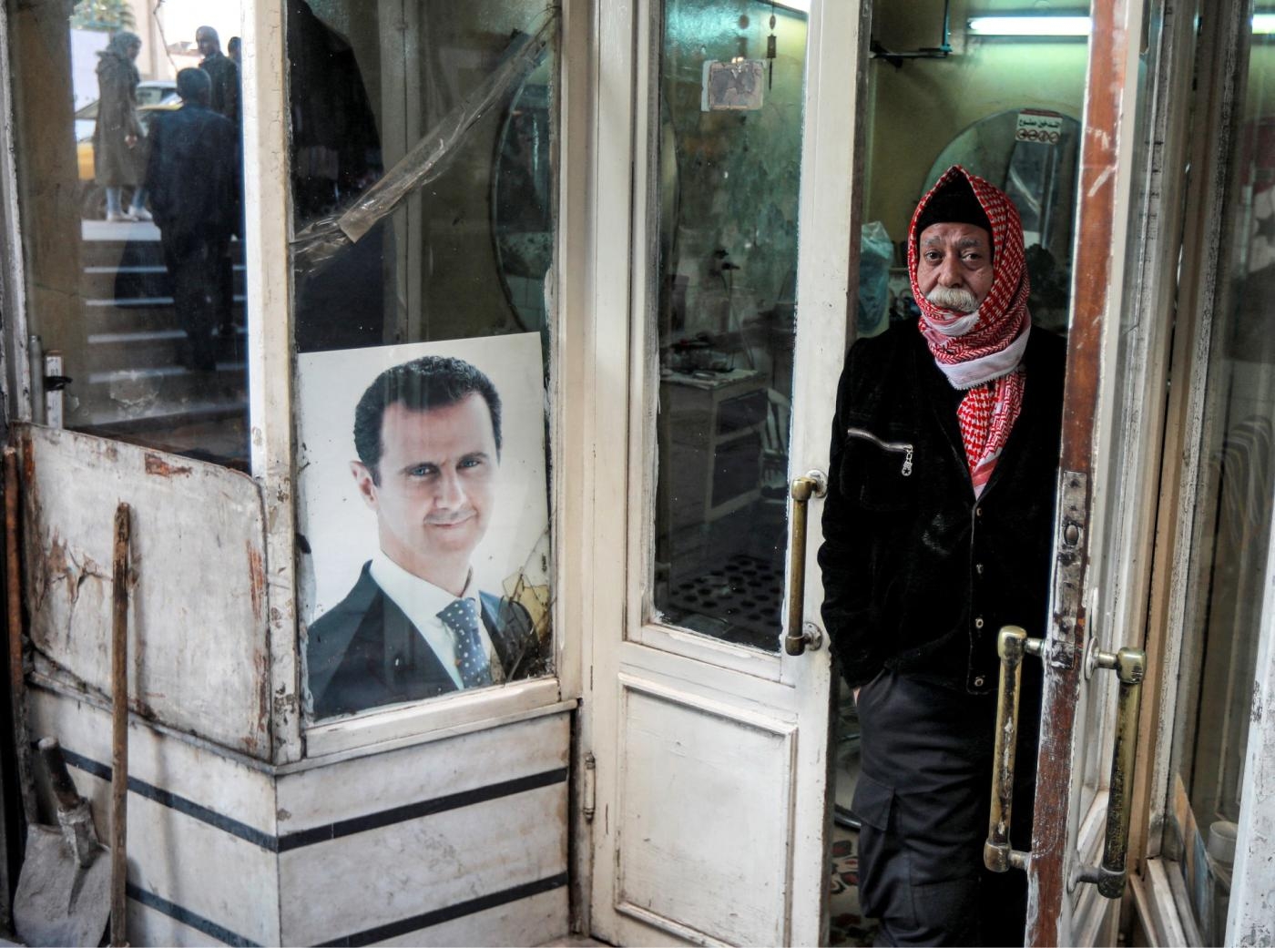 Un homme se tient à l’entrée d’un salon de coiffure à côté d’un portrait du président syrien Bachar al-Assad à Damas, le 15 décembre 2021 (AFP)