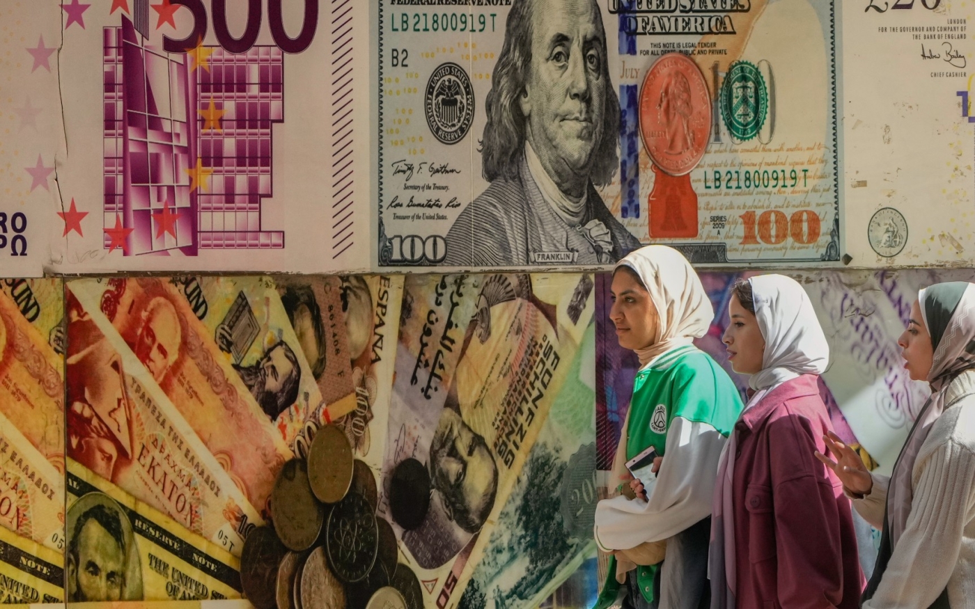 Des Égyptiennes passent devant un poster représentant le dollar américain et d’autres devises devant un bureau de change du Caire, le 12 janvier 2023 (AP)
