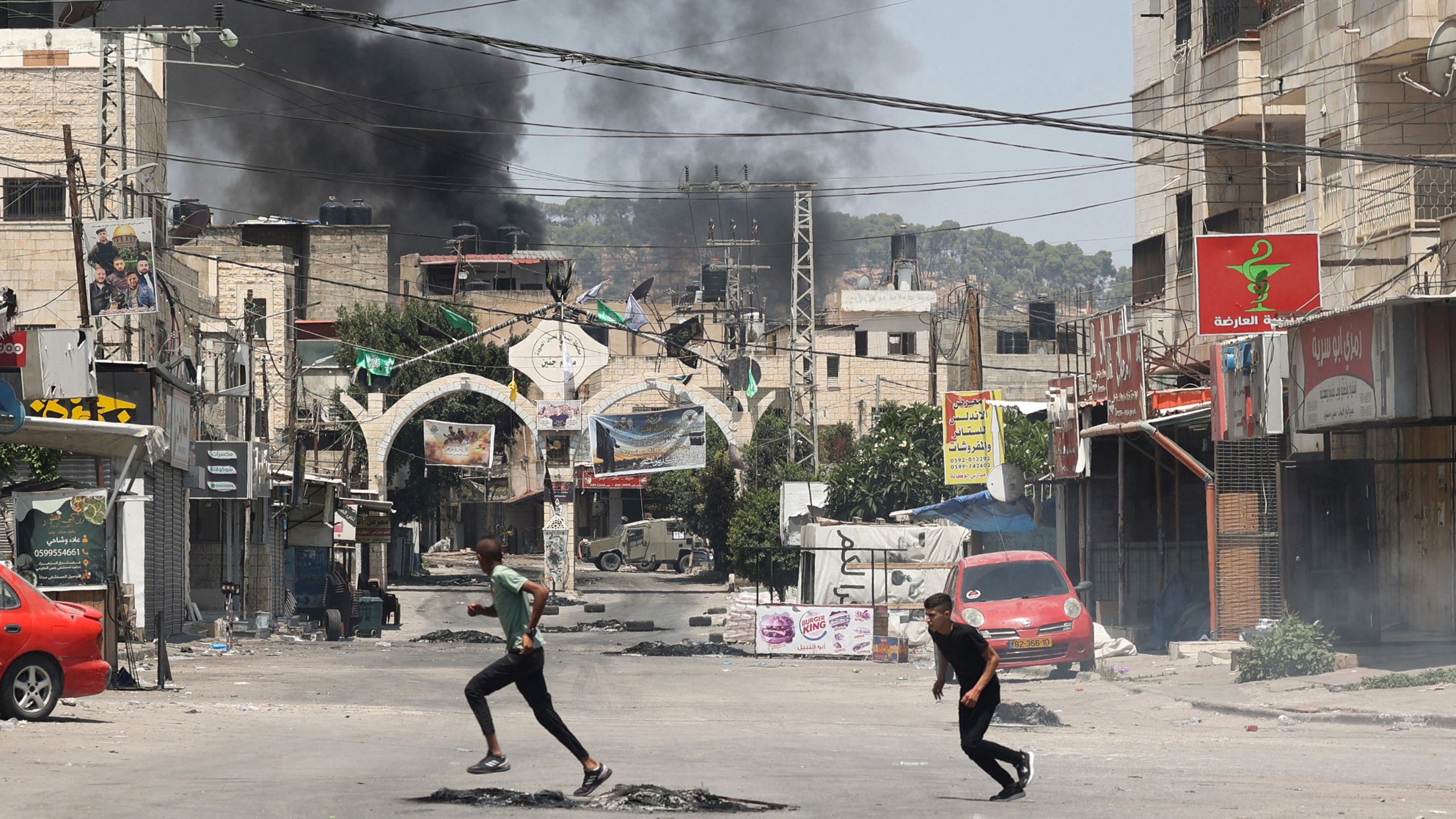 Palestinian youths run for cover near in Jenin as smoke rises amid an Israel offensive in the occupied West Bank city on 3 July 2023 (AFP)