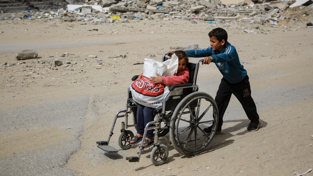 A boy pushes a young girl in a wheelchair past a destroyed building in Gaza City, on 28 March 2024 (AFP)
