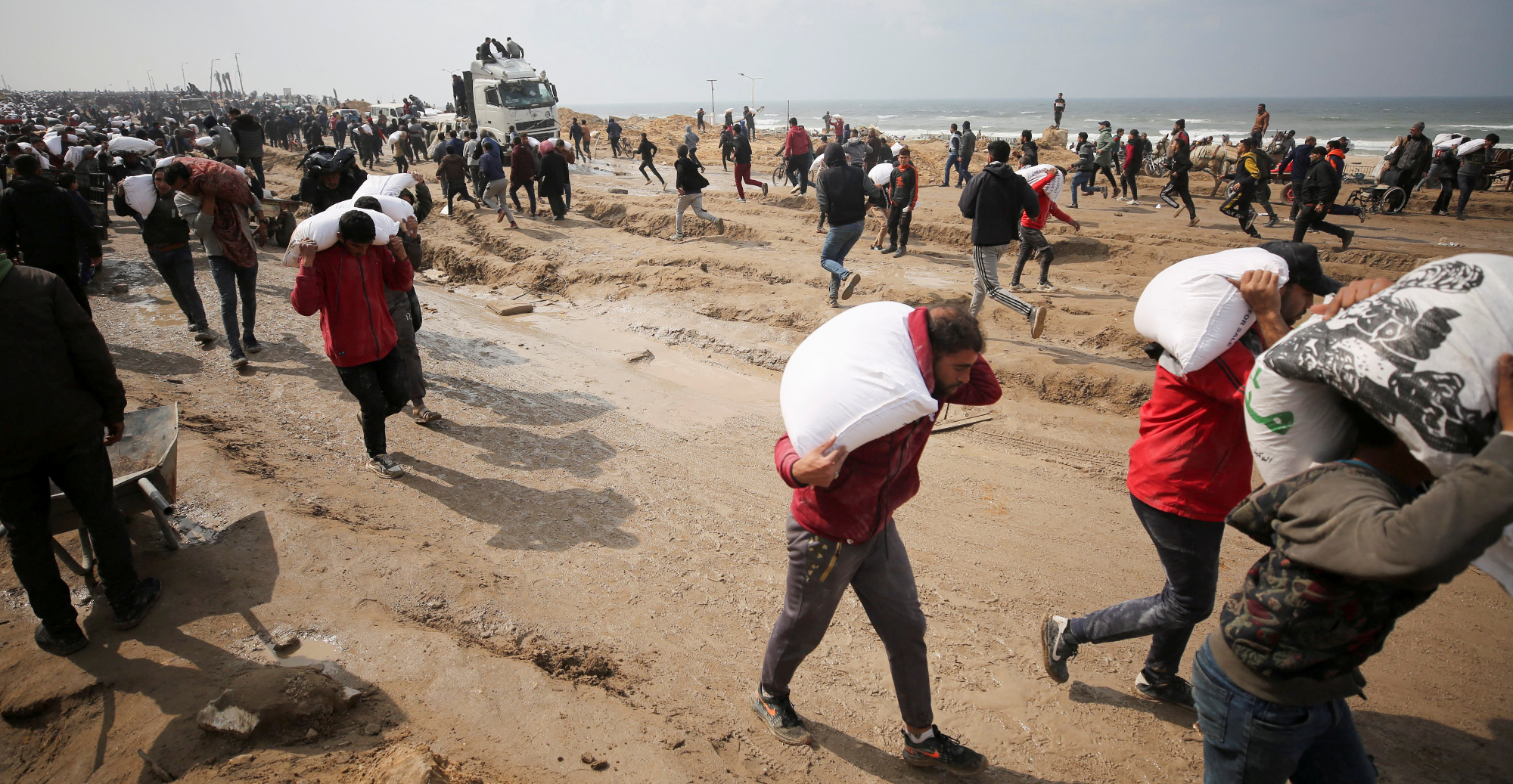 Palestinians carry bags of flour they grabbed from an aid truck near an Israeli checkpoint, as Gaza residents face crisis levels of hunger in Gaza City, 19 February 2024 (Reuters/Kosay Al Nemer)