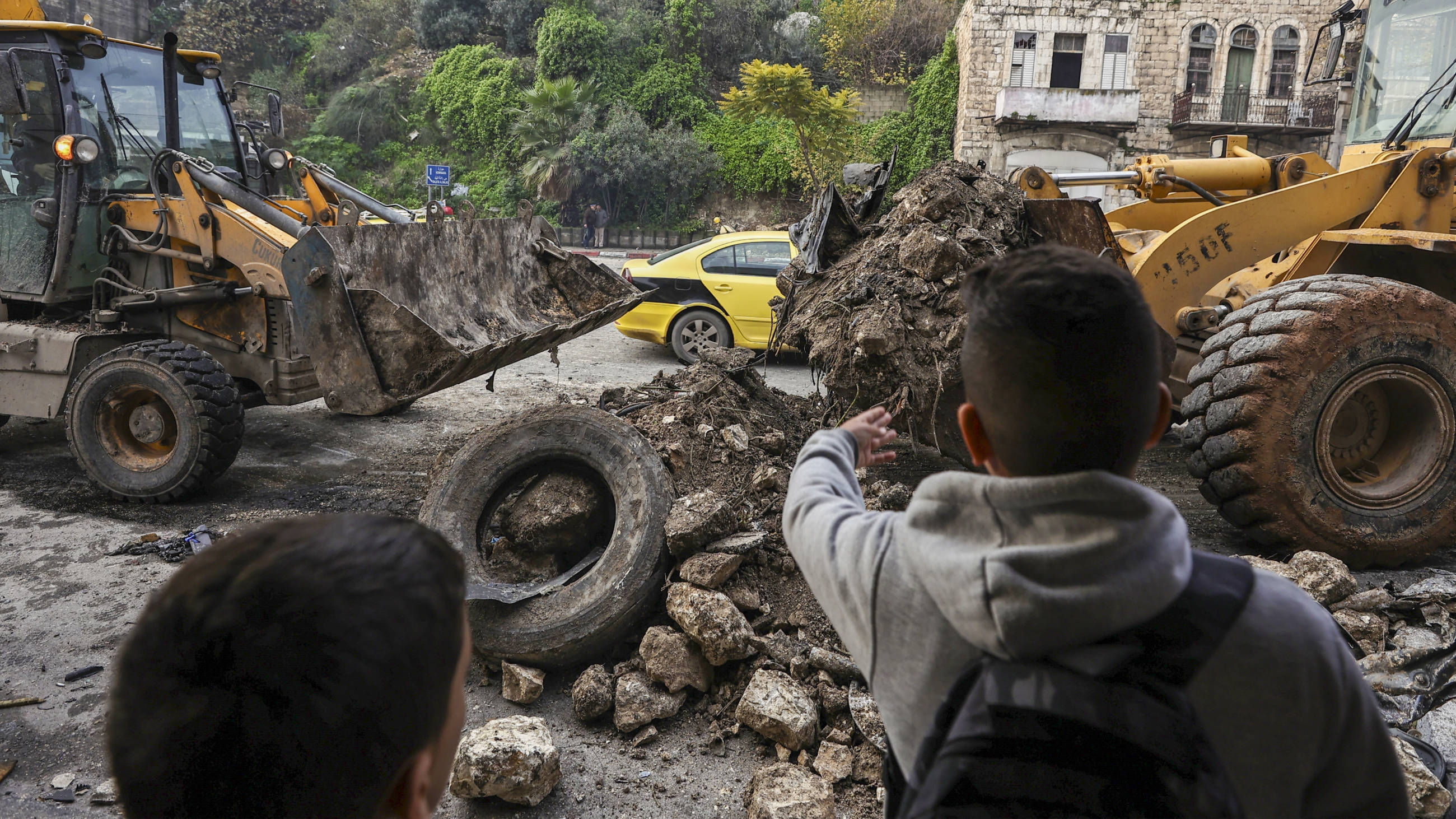 Children look on as bulldozers remove a destroyed car that was targeted in an Israeli airstrike in which five Palestinians were killed near the occupied West Bank Balata refugee camp, on 17 January 2024 (AFP)