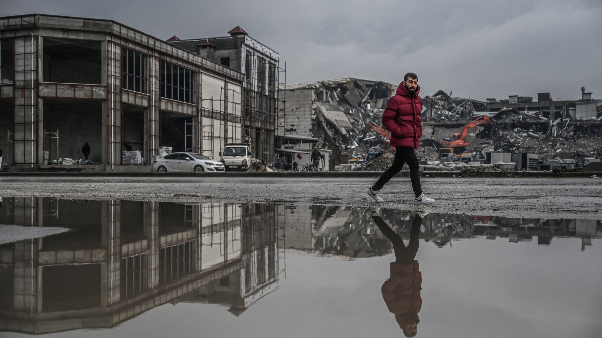 A man walks past a flooded street as diggers work to clean the rubble of collapsed buildings in Antakya 12 January (AFP/Ozan Kose)