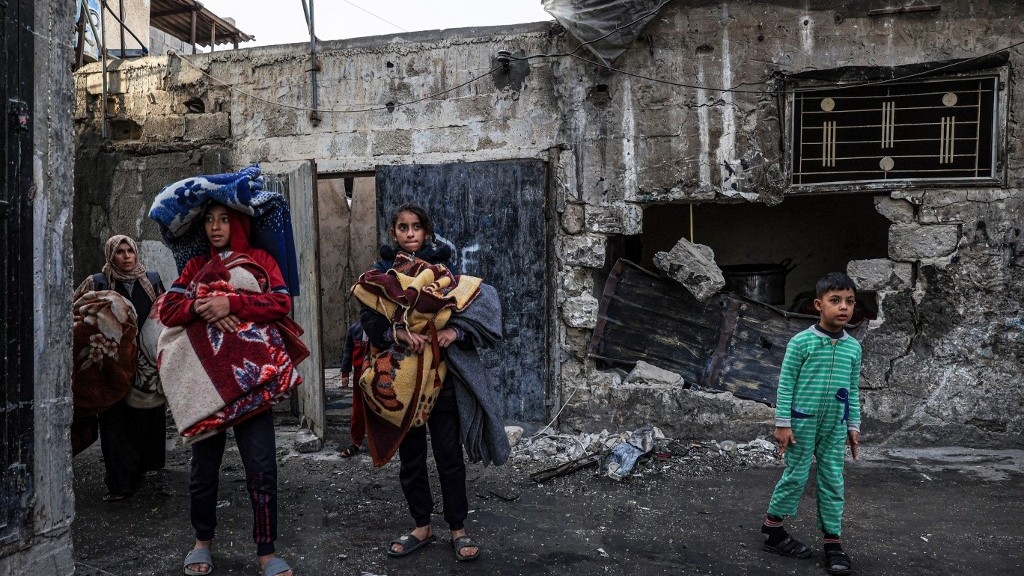 A woman and children carry blankets as they walk past houses destroyed in Israeli bombardments in Rafah, in the southern Gaza Strip, on 20 December 2023 (Said Khatib/AFP)
