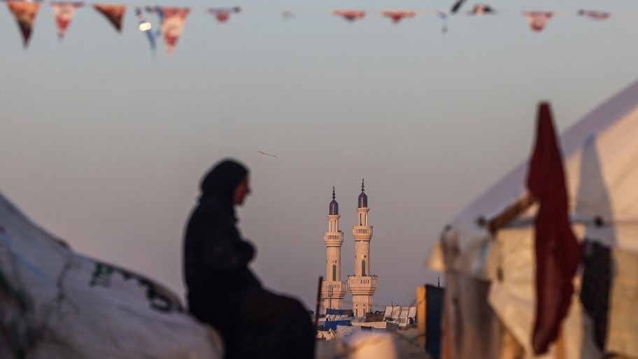 A Palestinian woman sits at a camp for displaced people backdropped by the minarets of a mosque, on the first day of Ramadan, in Rafah in the southern Gaza Strip on 11 March 2024 (AFP)