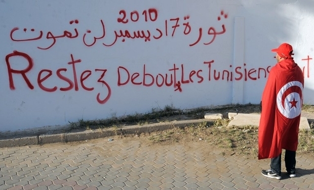 A Tunisian man draped in the national flag reads graffiti written in French, "Stay standing Tunisians", along Mohammed Bouazizi Avenue, in the impoverished central town of Sidi Bouzid, on 16 December 2013 (AFP)