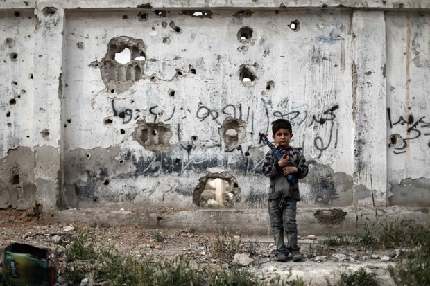 A Syrian boy stands with a toy gun in front of a bullet-riddled wall in the rebel-held town of Douma (AFP)